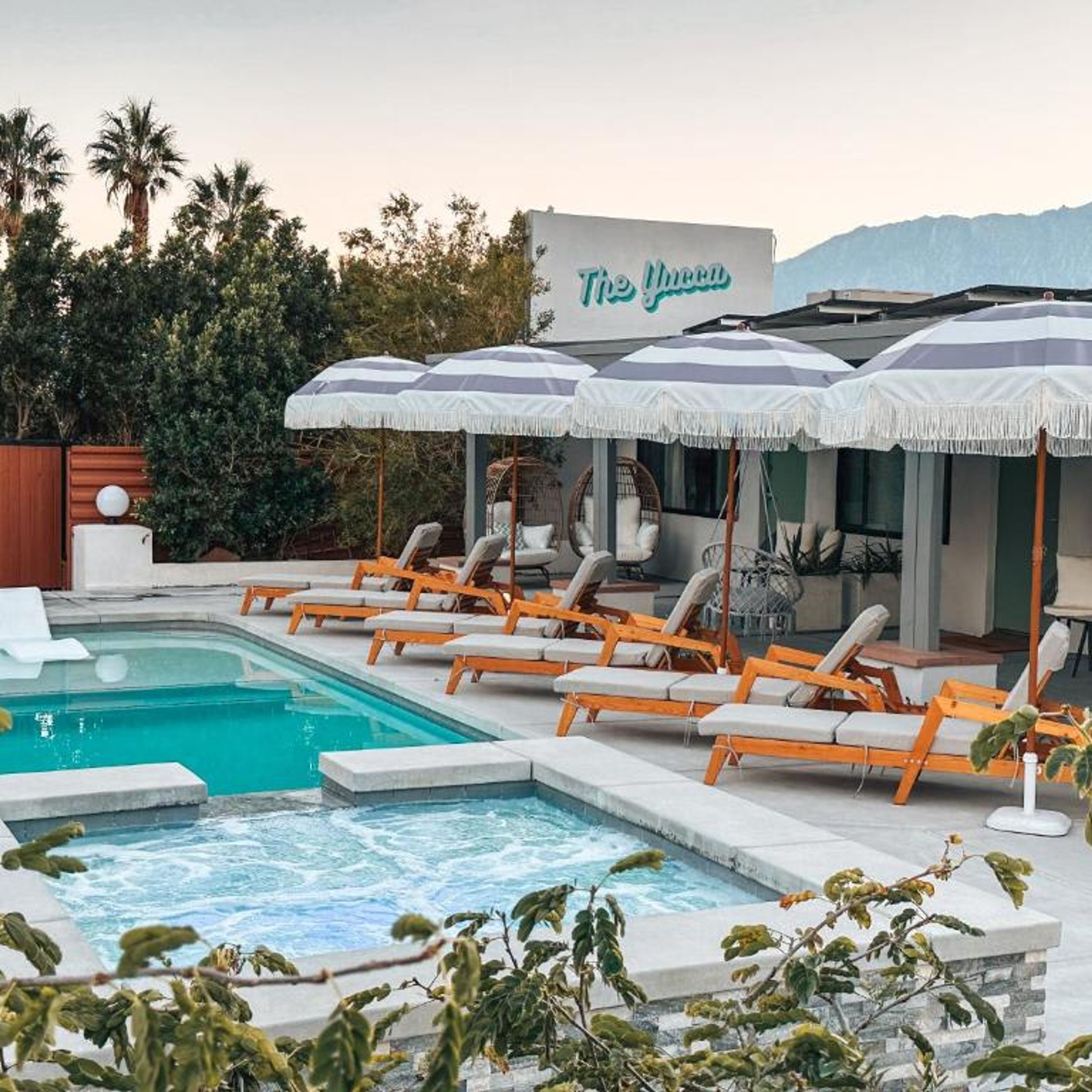 A pool area with lounge chairs and umbrellas, a sign reading "The Yuca", and a view of palm trees and mountains.