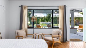 A modern hotel room with a large bed, two armchairs, a small table, and a view of a swimming pool and greenery outside. - The Yucca Hotel- Entire Boutique Buyout (Desert Hot Springs)