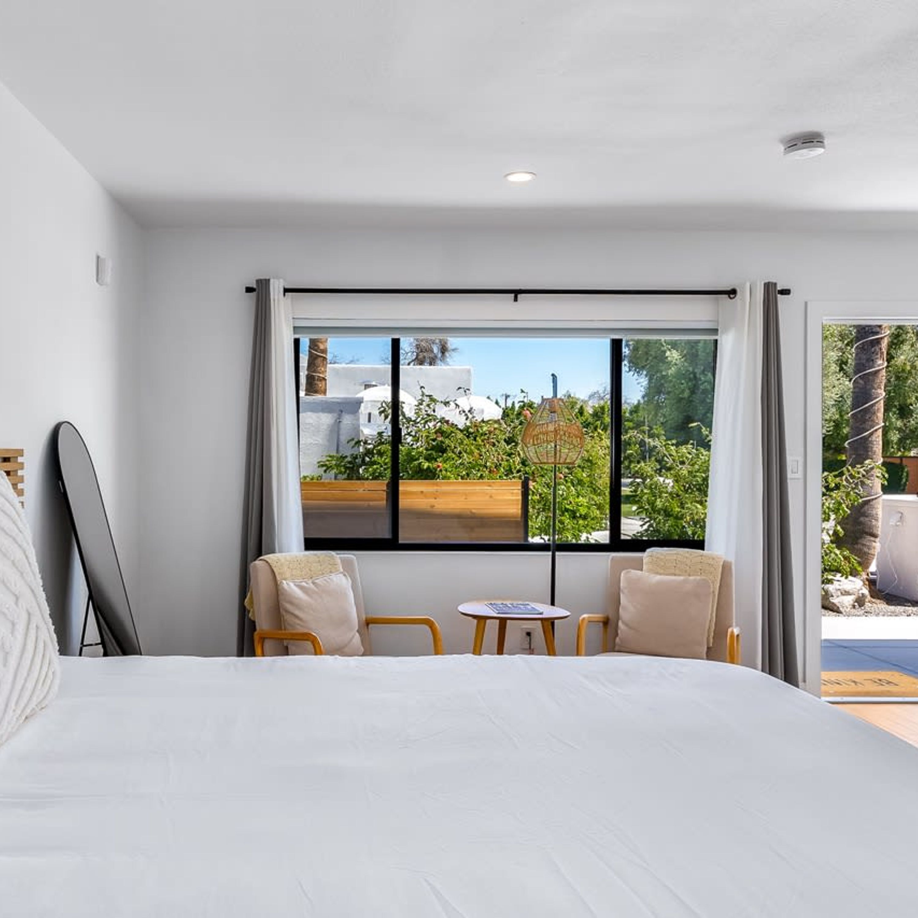 Ett modernt hotellrum med en stor säng, två stolar, ett litet bord och utsikt över en vit byggnad och grönområden utanför.