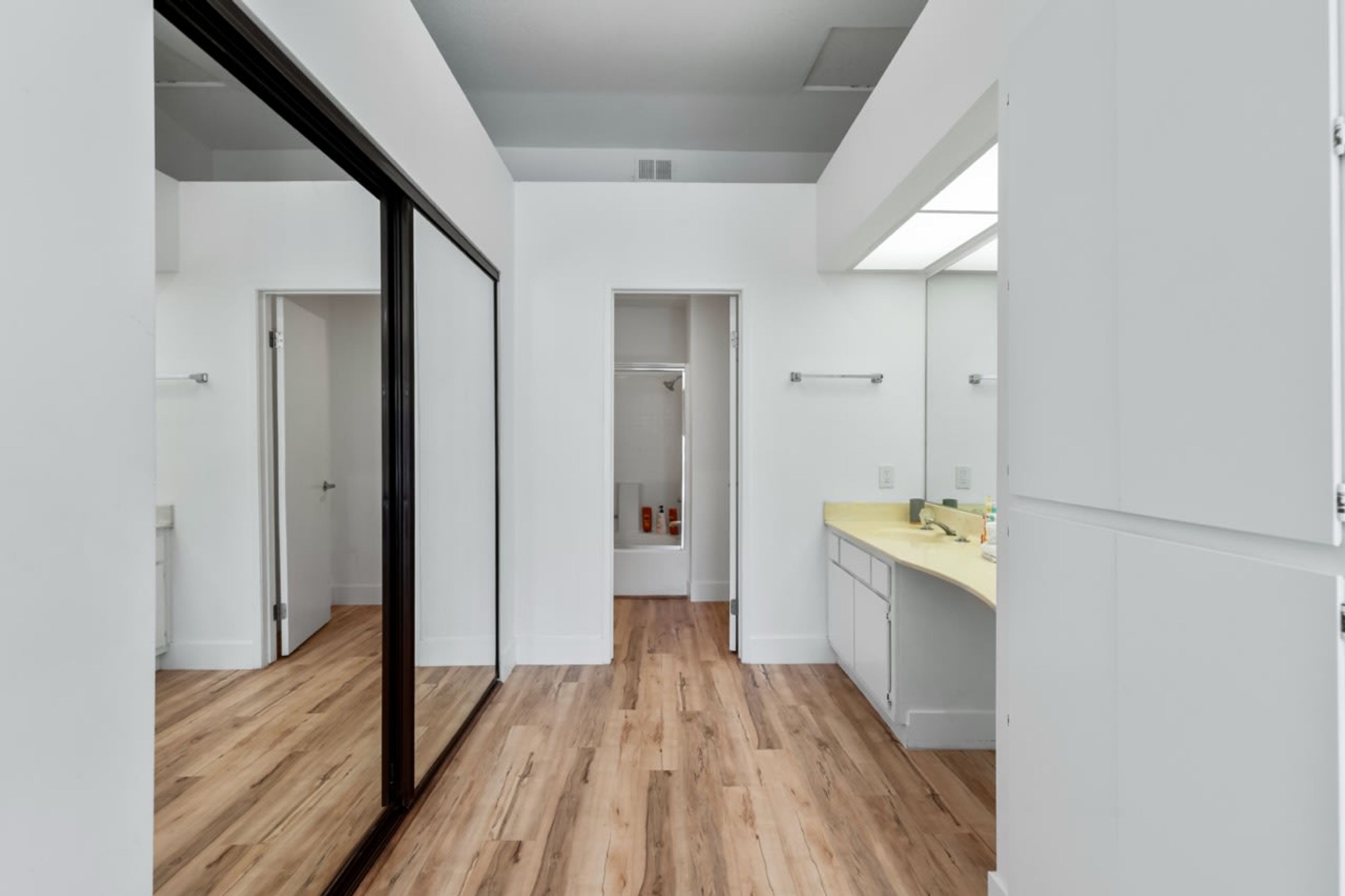 A modern, minimalist hallway with wooden flooring, white walls, and a glass door leading to a bathroom.
