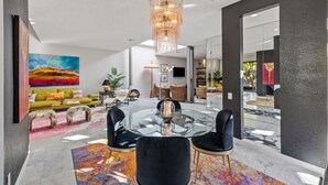 Ein modernes Wohnzimmer mit einem Glastisch, schwarzen Stühlen und einer Chandelier. Ein Sofa mit bunten Kissen, ein Gemälde an der Wand und ein Kamin sind ebenfalls vorhanden.