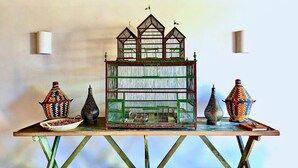 Una casa modelo con techo verde y varias ventanas, exhibida sobre una mesa con jarrones decorativos y una jaula pequeña para pájaros.