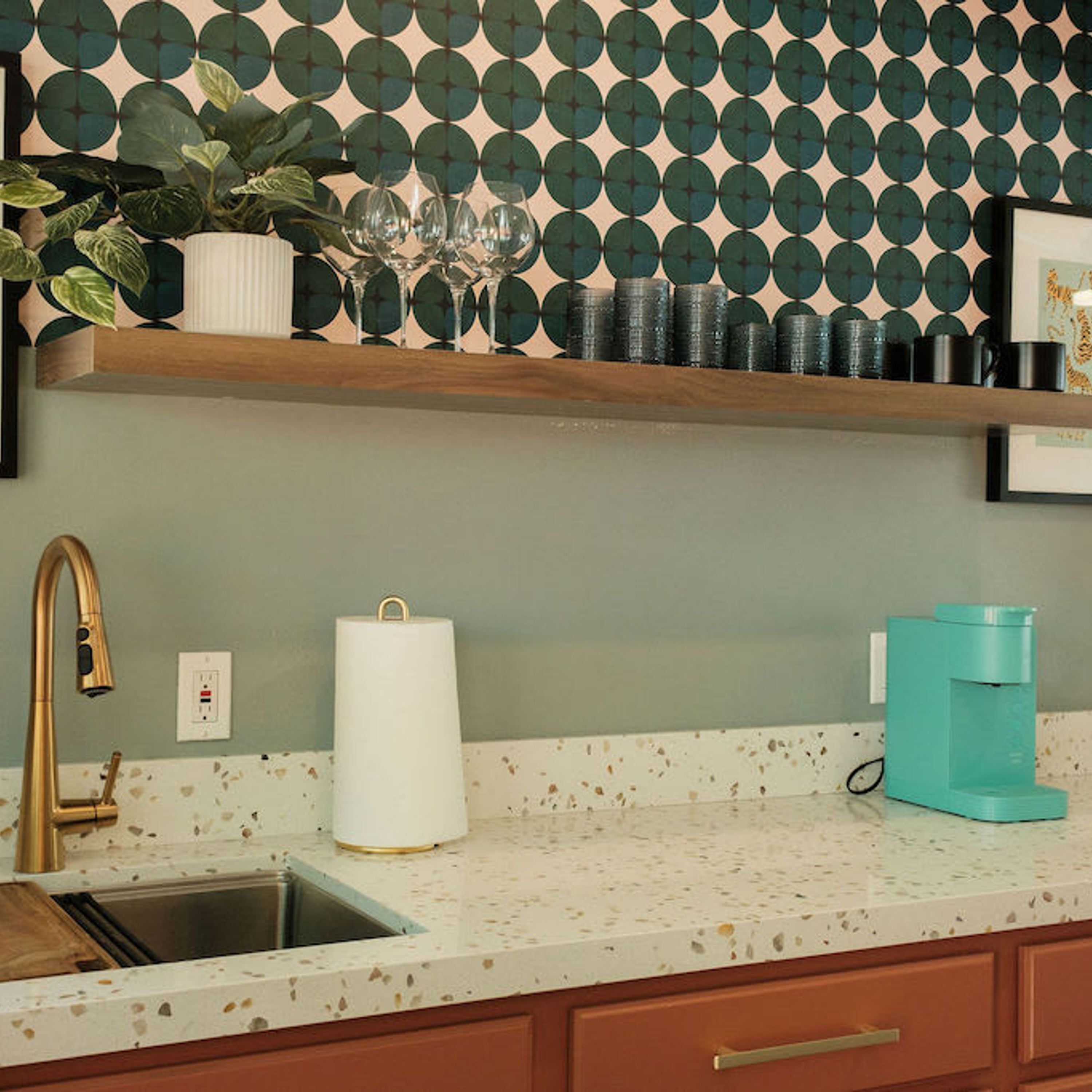 Une cuisine avec un plan de travail à motifs, un robinet doré, une cafetière turquoise et un mur décoré d’un motif.