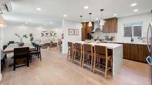 Eine moderne Küche mit Holzausstattung, einer zentralen Kochinsel mit Barhocker, Einbau-Deckenleuchten und einem Essbereich mit Holztisch und Stühlen.