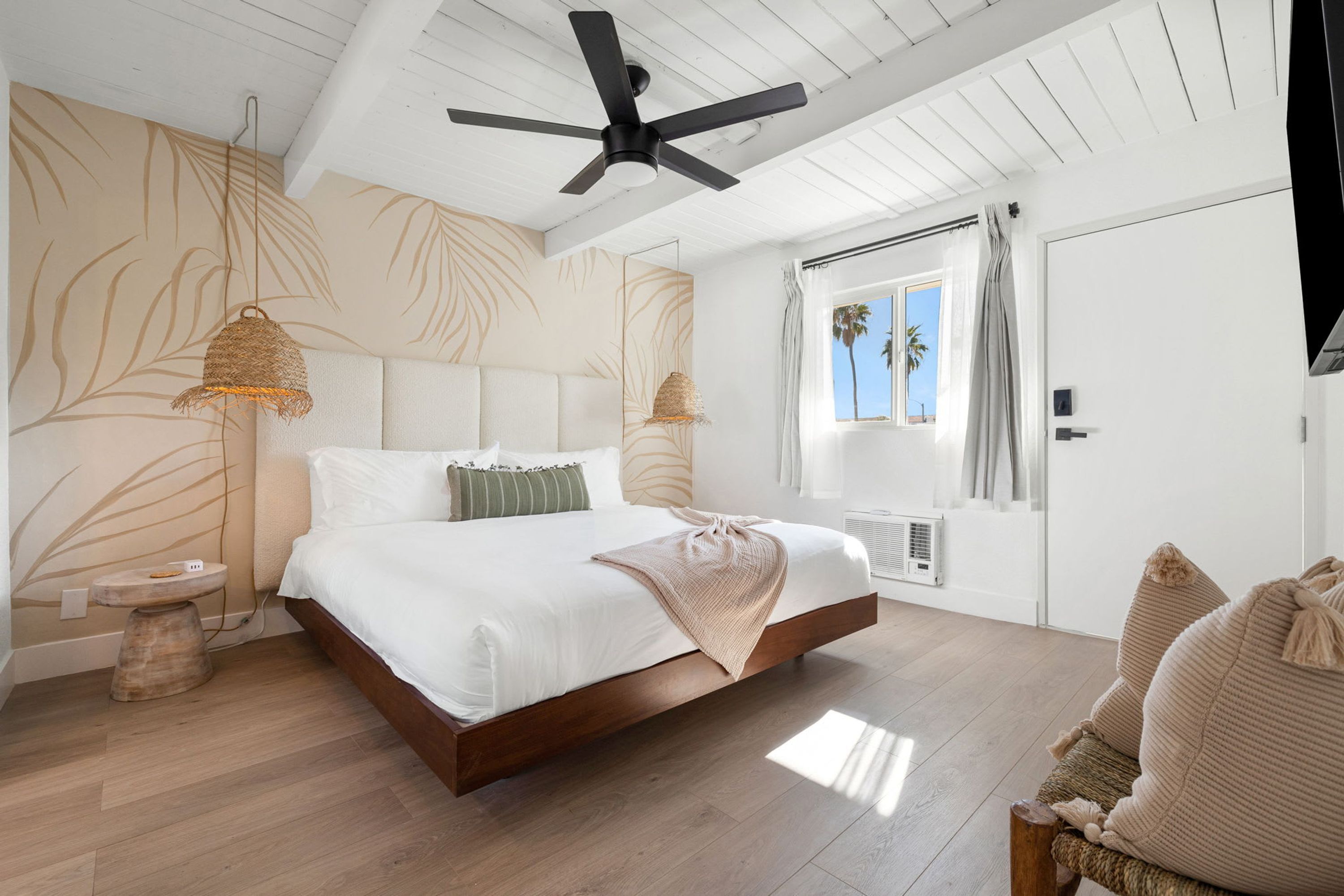 A bedroom with a bed, a ceiling fan, a window with curtains, and a view of palm trees.