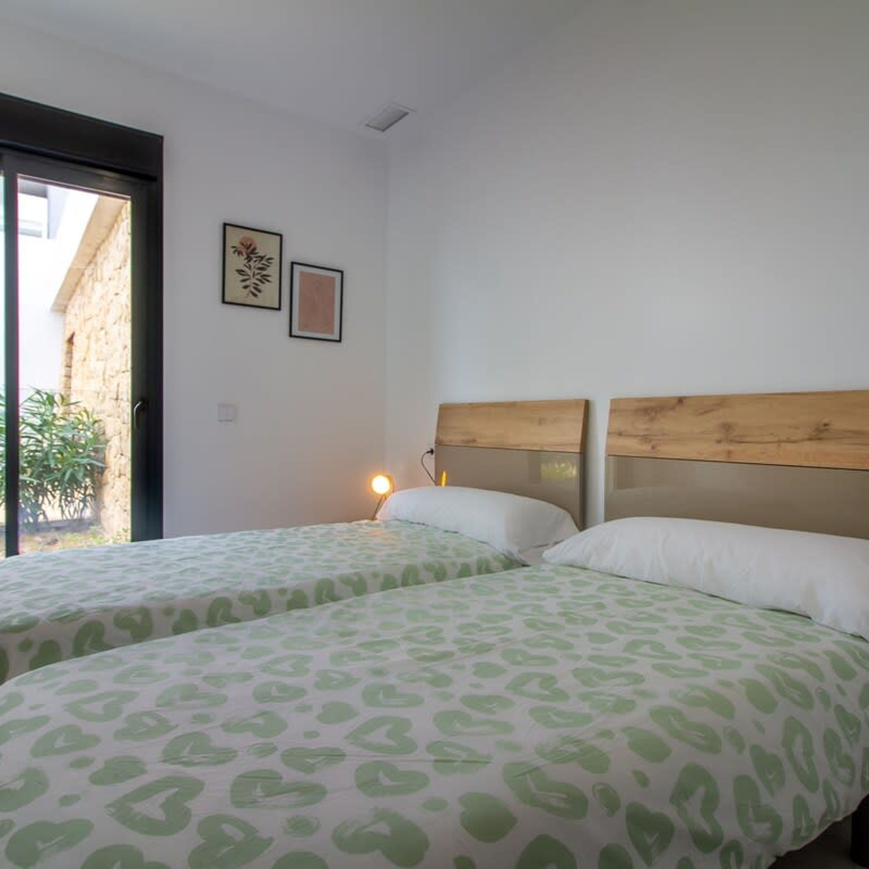 A room with two beds, each with a wooden headboard and green patterned bedding. There are two framed pictures on the wall, and a window with a view of greenery outside.