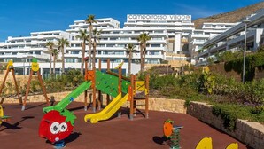 A playground with colorful play structures and a building labeled "Camorrosso Village" in the background. - Camporrosso 1C by Bookaparadise (Finestrat)