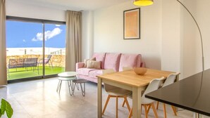 A modern living room with a wooden dining table, a pink sofa, and a large window offering a view of a green outdoor area. - Camporrosso 1C by Bookaparadise (Finestrat)