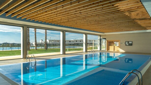A modern indoor swimming pool with clear glass walls, wooden ceiling beams, and a view of a cityscape. - Camporrosso 1C by Bookaparadise (Finestrat)