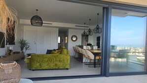 Un espacio habitable moderno con un sofá verde, un área de comedor y un gran ventanal que da a un balcón con vistas a la ciudad.