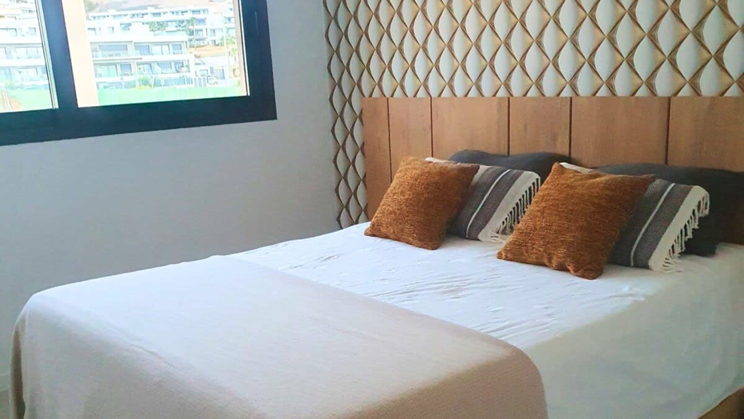 A bed with a wooden headboard and multiple pillows, a window with a view of buildings, and a plain wall.