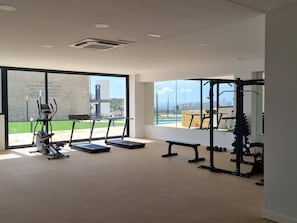 A spacious gym with treadmills, a bench, and a squat rack, featuring large windows with a view of a pool and buildings. - Casa la Vista by Bookaparadise (Finestrat)