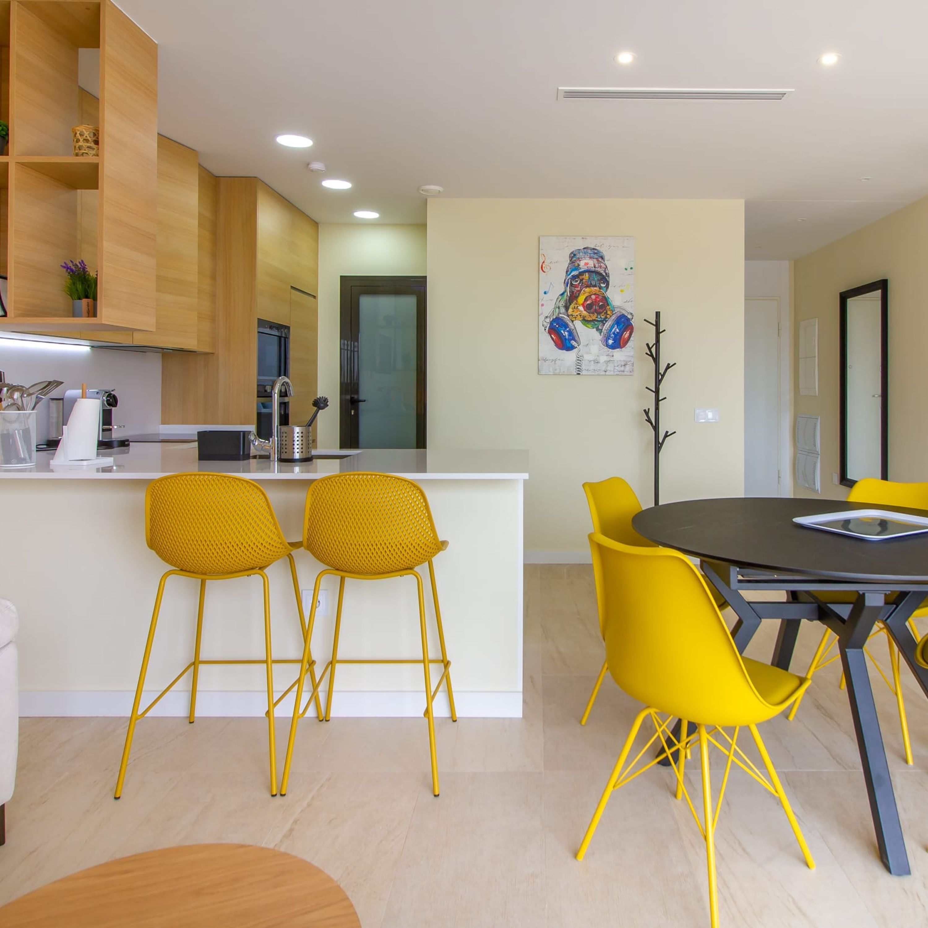 A modern kitchen with wooden cabinets, a built-in oven, and a sink. There are two yellow chairs and a round table with a tablet.
