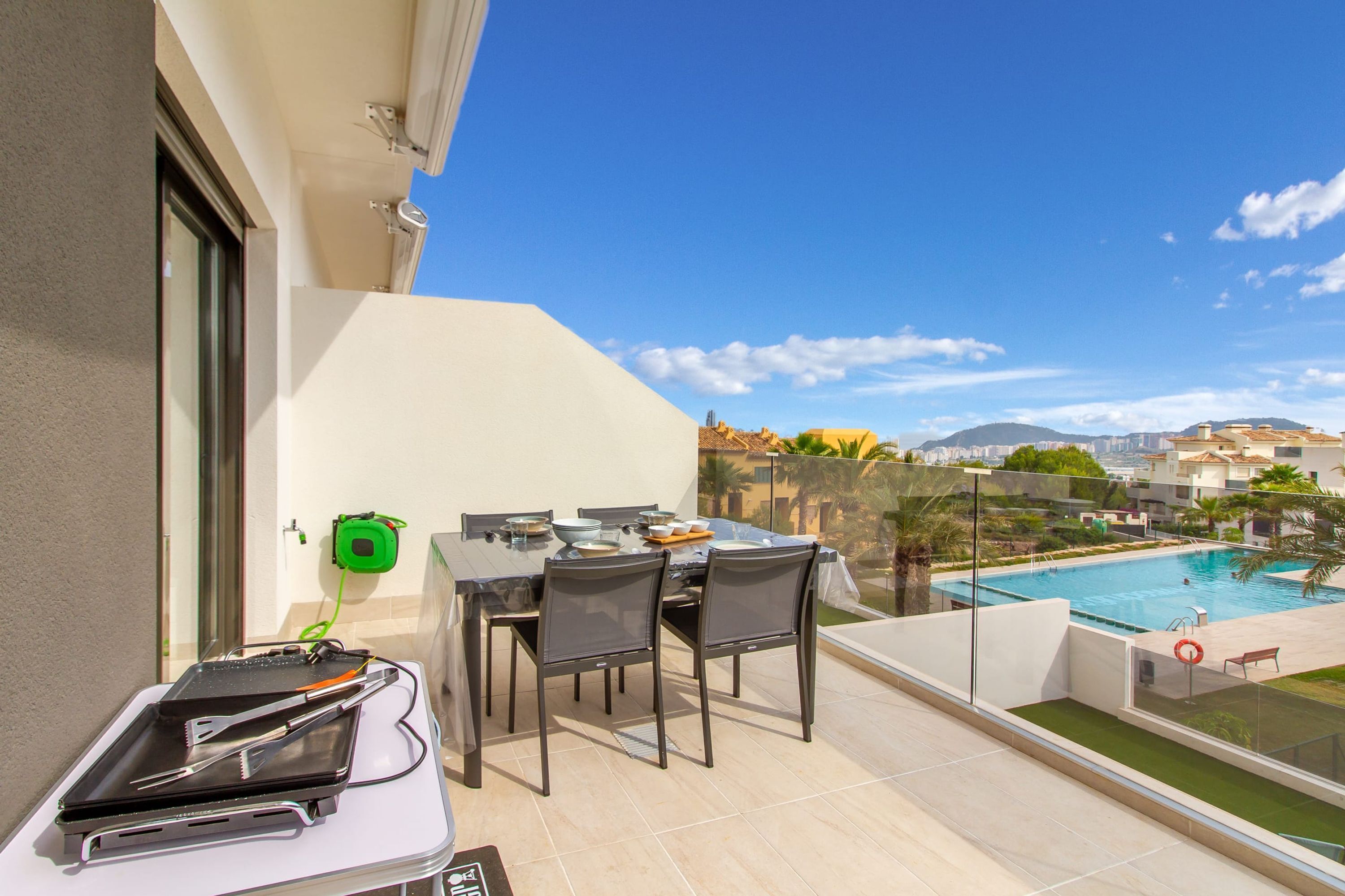 Varanda com mesa de jantar posta para quatro, com vista para uma piscina e a paisagem urbana.