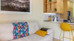 A modern living room with a sofa, a colorful throw pillow, and a yellow cushion. There is a kitchen area with wooden cabinets and a bar stool. - Mediterranean Views by Bookaparadise (Finestrat)