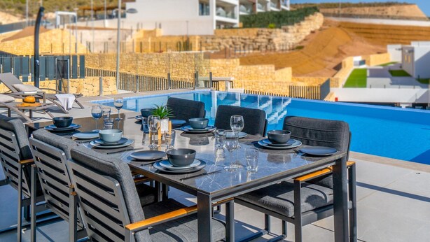 A dining area with a pool view, outdoor seating, and a modern building in the background. - Villa Esmeralda 20 by Bookaparadise (Finestrat)