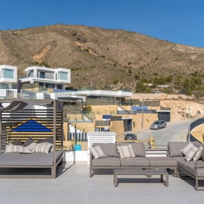 Un espacio exterior moderno con zona de estar, sofá y estructura de madera, con vistas a un paisaje montañoso y edificios contemporáneos.