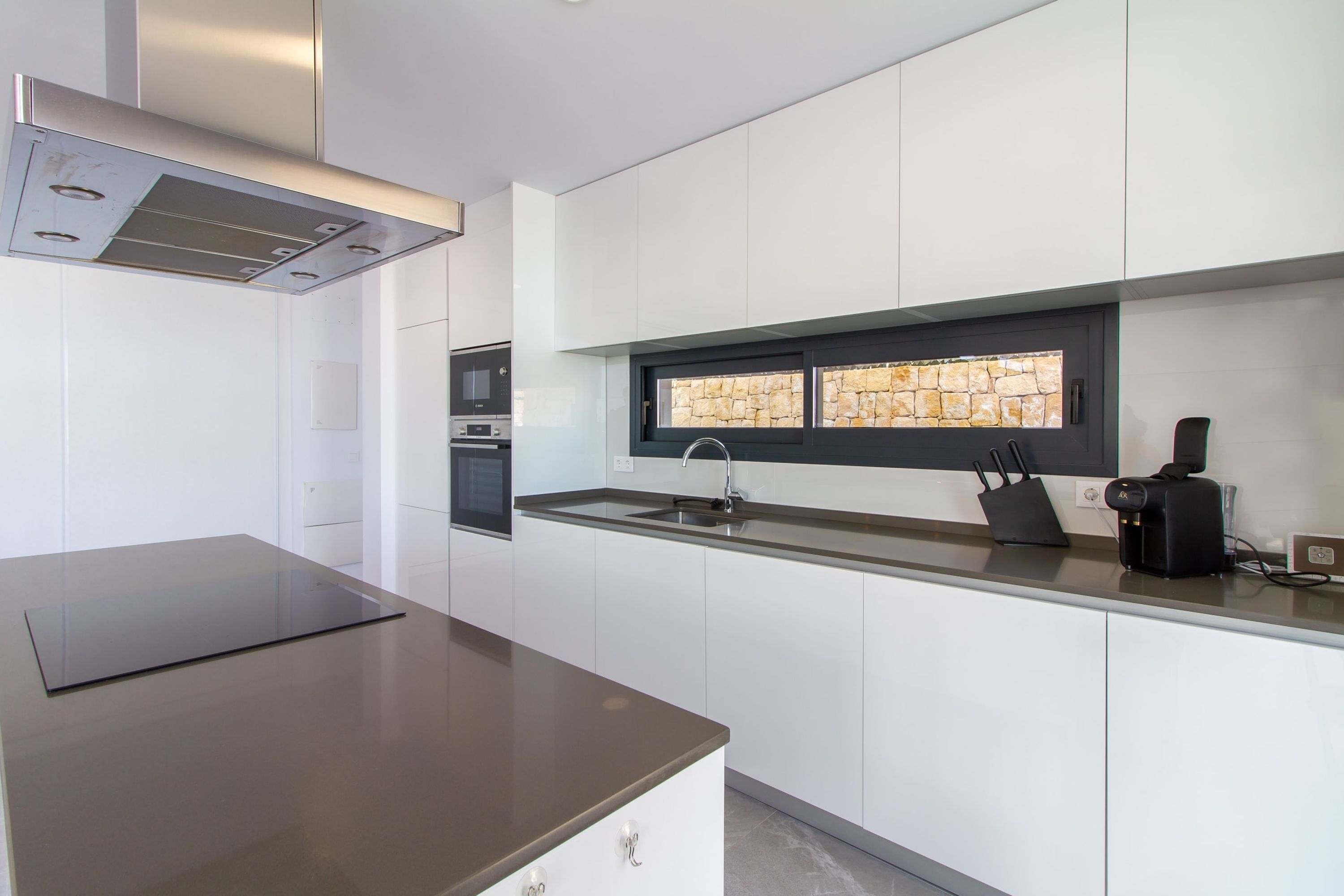 A modern kitchen with white cabinets, a stainless steel countertop, a built-in oven, and a window with a stone backsplash.