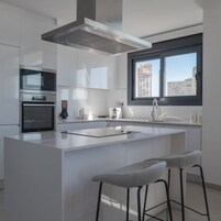 Una cocina moderna con armarios blancos, una isla central con taburetes, una campana extractora de acero inoxidable y un ventanal con vista a la ciudad.