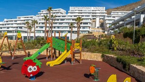 Une aire de jeux avec des équipements colorés et des palmiers devant un bâtiment blanc doté de balcons.