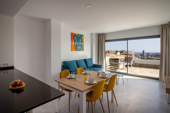 A modern living space with a dining area, a sofa, and a large window offering a city view. - Camporrosso Blue Sea by Bookaparadise (Finestrat)