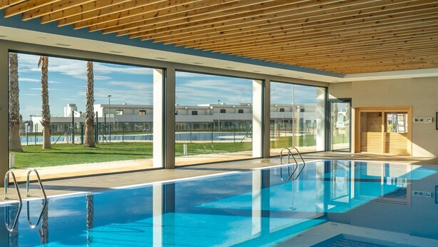 Une piscine intérieure avec des parois en verre transparent, un plafond en bois et une vue sur un ensemble de bâtiments à l’extérieur.
