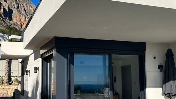 A modern outdoor dining area with a glass door leading to a covered space, a stone wall, and a mountain view. - Polop Hills 21A by Bookaparadise (Polop)