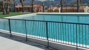 A rectangular swimming pool with a black metal railing, surrounded by palm trees and buildings with red roofs, set against a mountainous backdrop. - Polop Hills 21A by Bookaparadise (Polop)