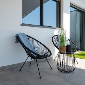 Een modern buitenzitgedeelte met twee zwarte frame stoelen, waarvan er een bekleed is met een denim jasje. Er staat een klein rond tafeltje met een gevlochten mand en een potplant erop.