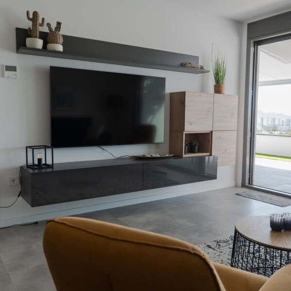 A modern living room with a flat-screen TV mounted on the wall, a wooden cabinet with a plant, and a large window providing a view outside.