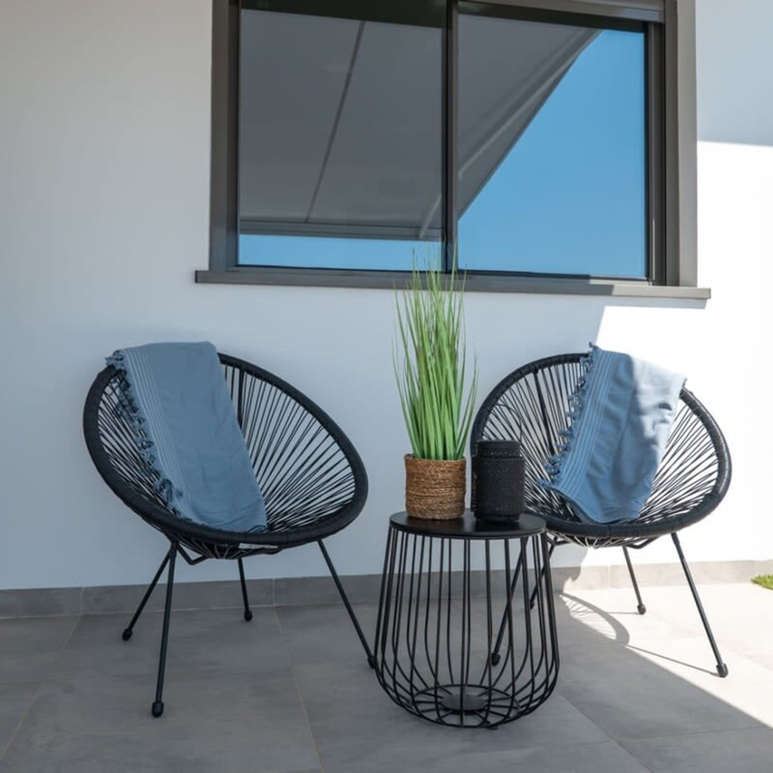 Dos sillas de estructura metálica negra con cojines azules, una mesita redonda con una cesta trenzada y una planta en maceta, y un ventanal grande con toldo blanco.