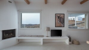 A modern living room with a fireplace, a bench, a TV, and large windows offering views of the outdoors. - Lea Villa 2 Mykonos (Mykonos Island)