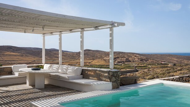 A white outdoor seating area with a pool, stone walls, and a view of the landscape. - Lea Villa 2 Mykonos (Mykonos Island)