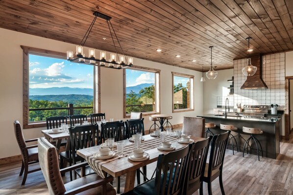 Un coin repas avec une table en bois dressée pour six personnes, de grandes fenêtres offrant une vue sur les montagnes et une cuisine au design moderne.