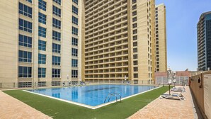 A rooftop swimming pool with a clear blue pool, surrounded by lounge chairs and umbrellas, set against a backdrop of high-rise buildings. - WelHome - Greenery Apt for Modern Studio in Production City (Dubai)