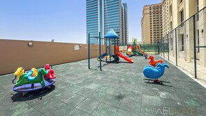 A playground with various play structures, surrounded by tall buildings and a chain-link fence. - WelHome - Greenery Apt for Modern Studio in Production City (Dubai)