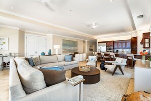 A spacious living room with a large sofa, a coffee table, and a dining area with chairs and a kitchen in the background. - Luxury Penthouse Condo With 360 Oceanviews (Seven Mile Beach)