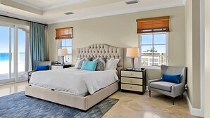 A spacious bedroom with a large bed, two bedside tables, a chair, a ceiling fan, and a view of the ocean. - Luxury Penthouse Condo With 360 Oceanviews (Seven Mile Beach)
