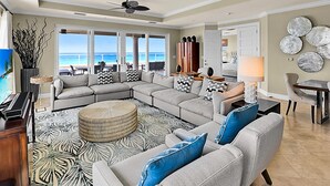 A spacious living room with a large sectional sofa, a round ottoman, and a view of the ocean through large windows. - Luxury Penthouse Condo With 360 Oceanviews (Seven Mile Beach)