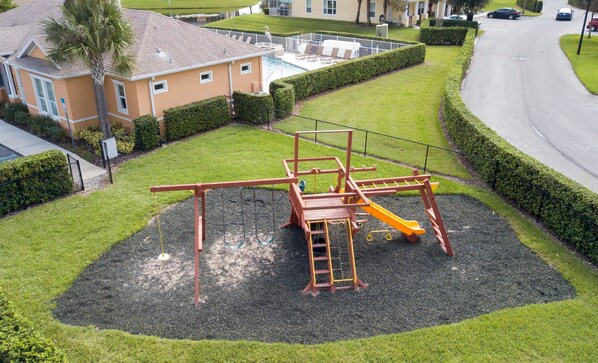 A playground with a slide and swings, surrounded by a well-maintained lawn and a building in the background. - Charming 3Bd w Pool Close to Disney Serenity 1639 (Clermont)
