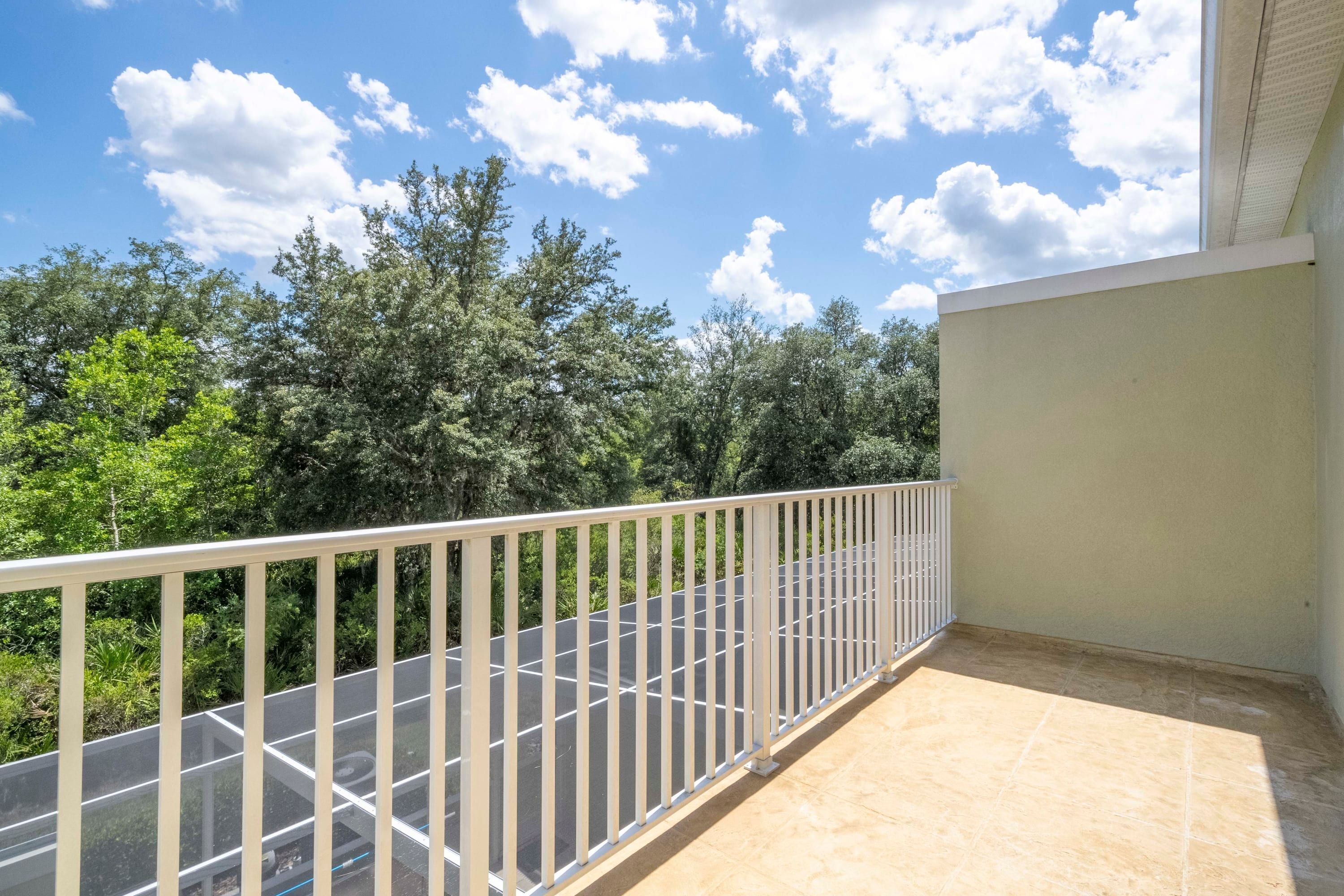 A balcony with a metal railing, a tiled floor, and a view of trees and a clear sky.
