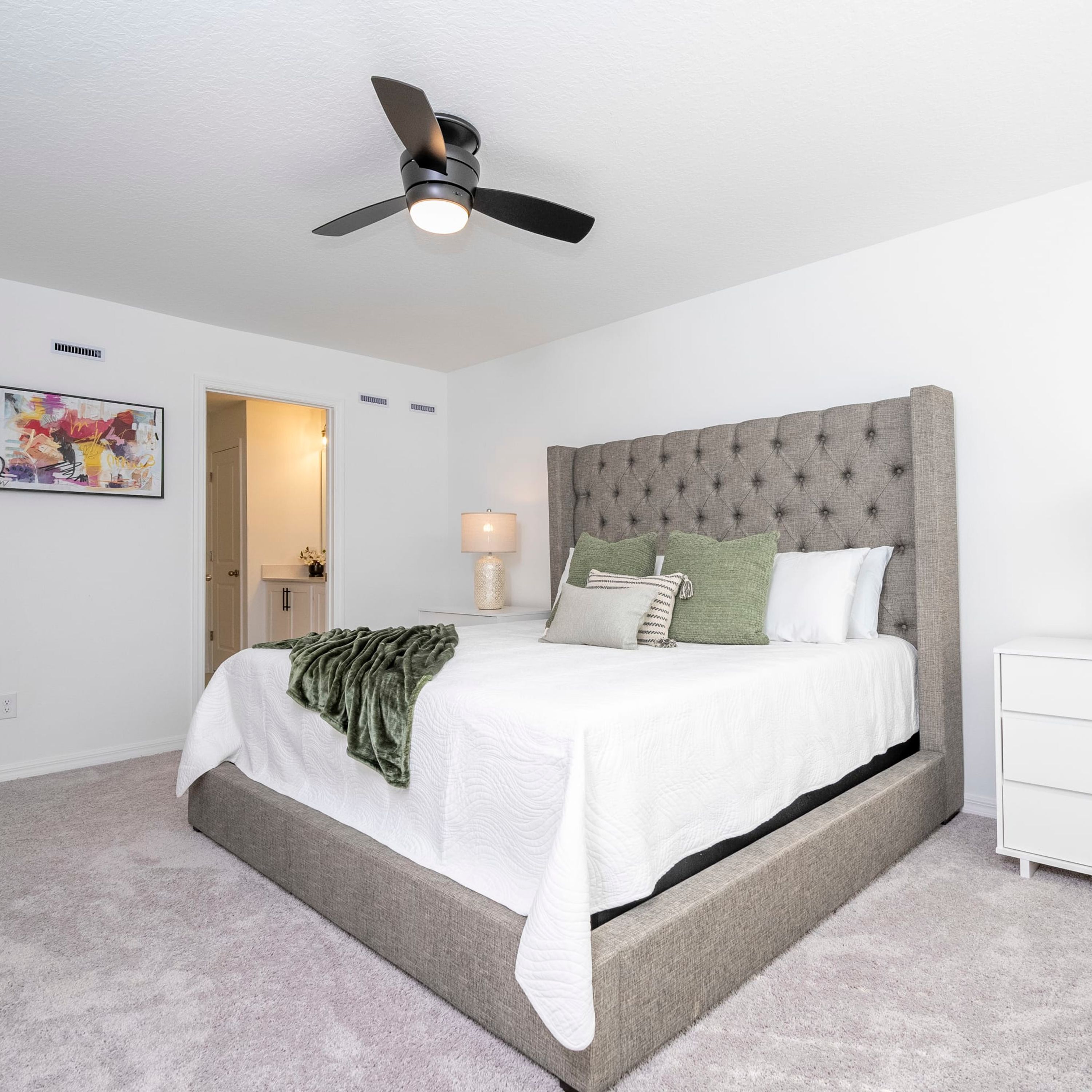 Un dormitorio con cama, ventilador de techo, mesita de noche, lámpara y puerta que da a otra habitación.