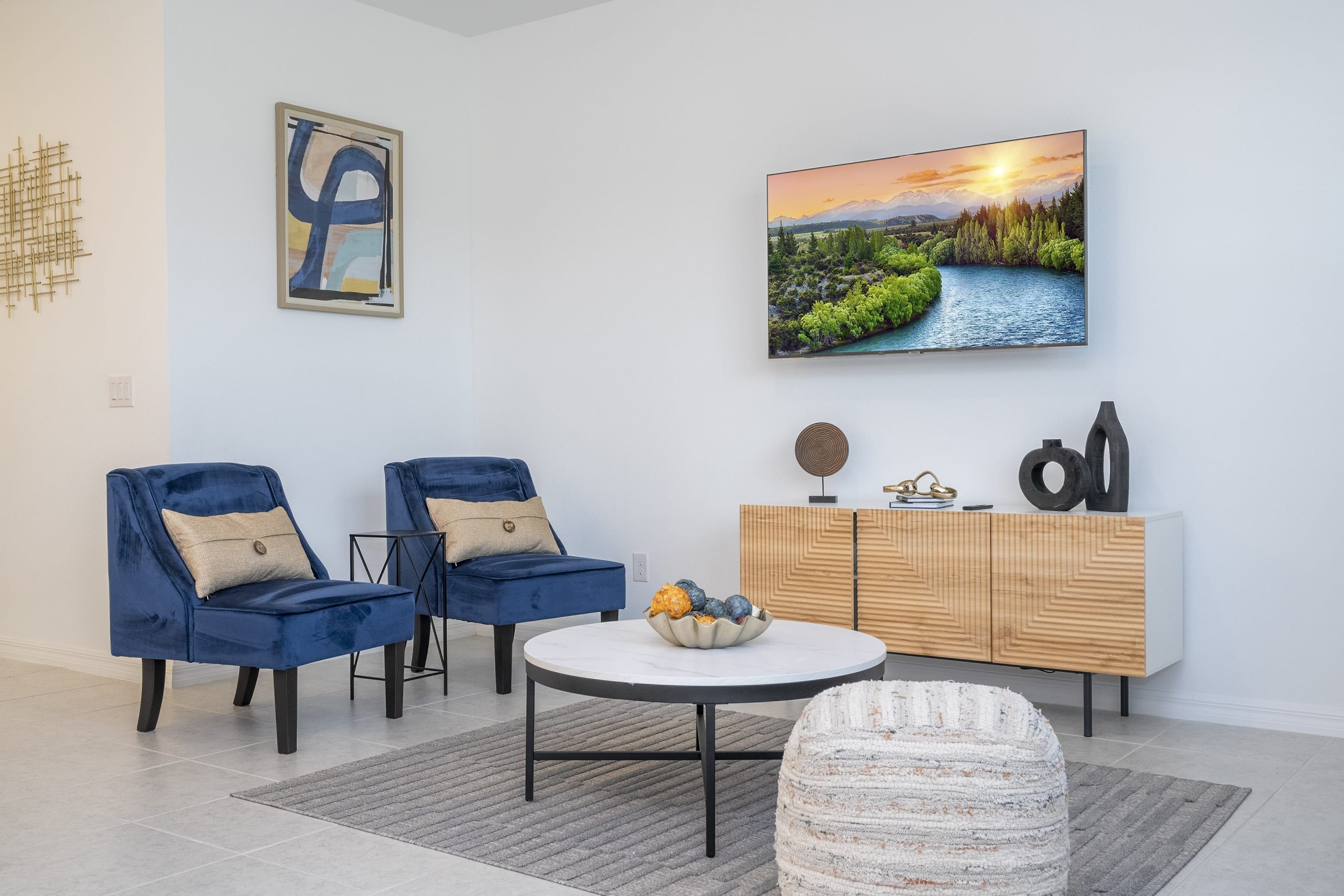 A modern living room with two blue armchairs, a round coffee table, a white ottoman, a wooden cabinet with decorative items, and a landscape painting on the wall.