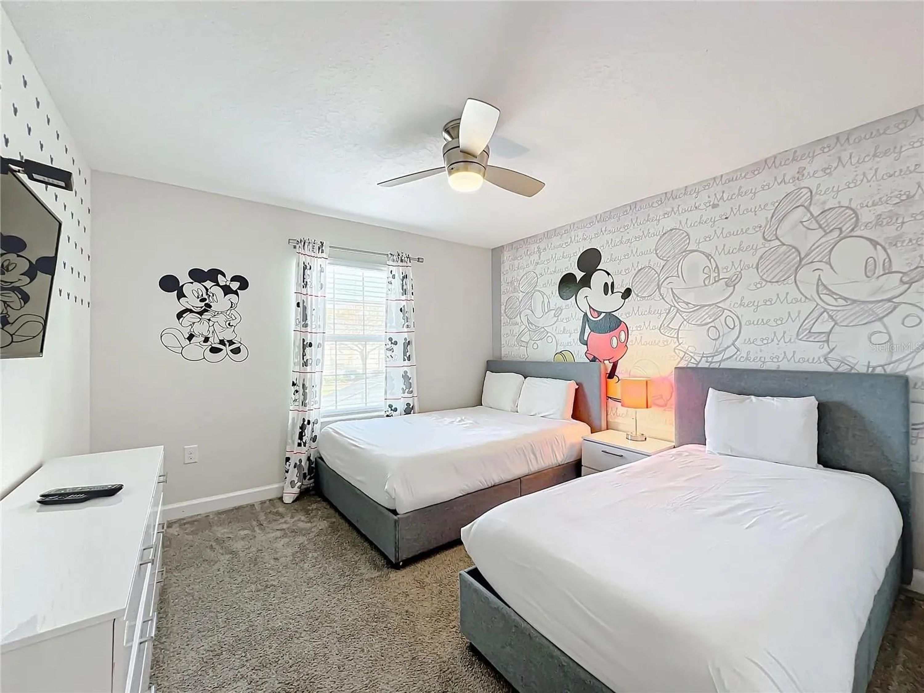 Une chambre d’hôtel avec deux lits, un ventilateur de plafond, des autocollants de Mickey Mouse sur les murs et une fenêtre avec des rideaux.
