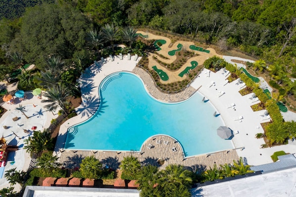 Eine Vogelperspektive zeigt ein Resort mit einem großen Schwimmbecken, Liegestühlen und einem angelegten Garten.