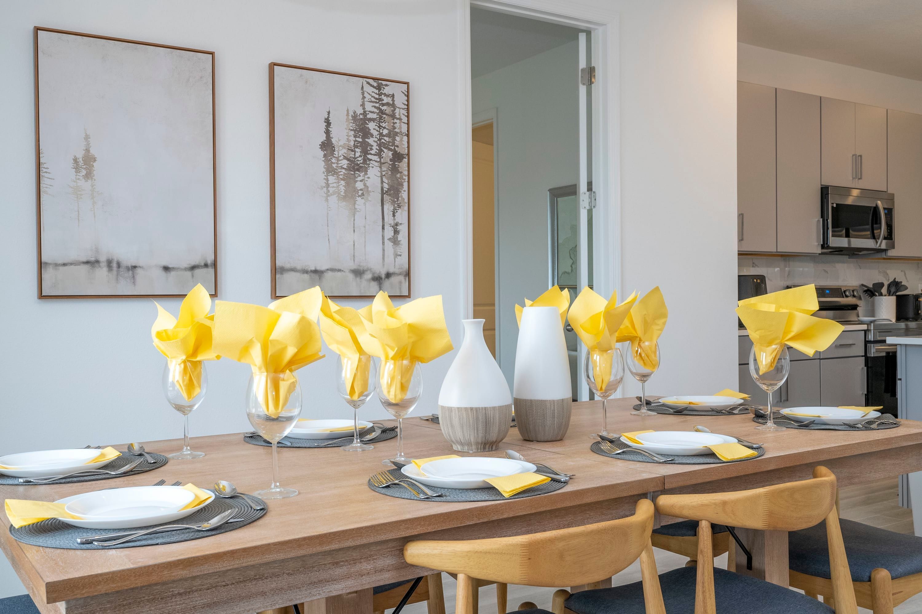 A dining table set with yellow napkins, white plates, and silverware, flanked by two framed pictures on the wall.