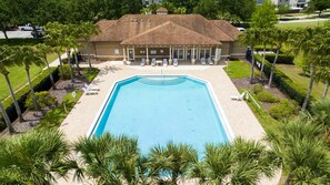 Ein großer rechteckiger Pool mit einer gefliesten Terrasse darum, eine überdachte Terrasse mit Sitzgelegenheiten im Freien und ein Gebäude mit Backsteinfassade im Hintergrund.