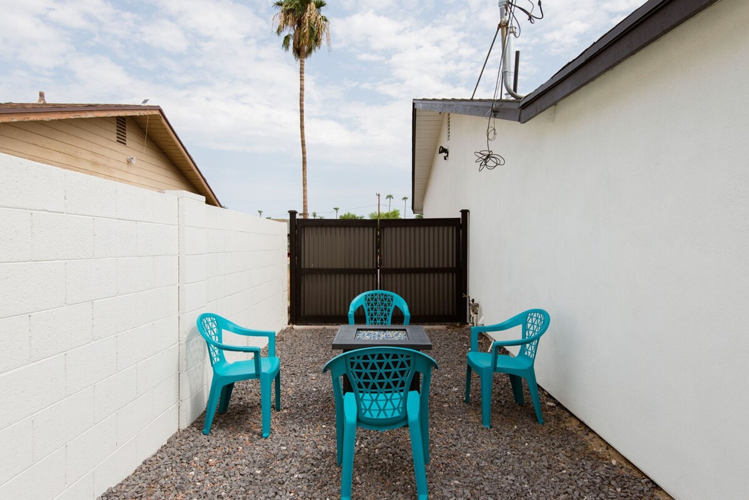 A small outdoor patio with turquoise plastic chairs and a table, surrounded by a white wall and a wooden roof.