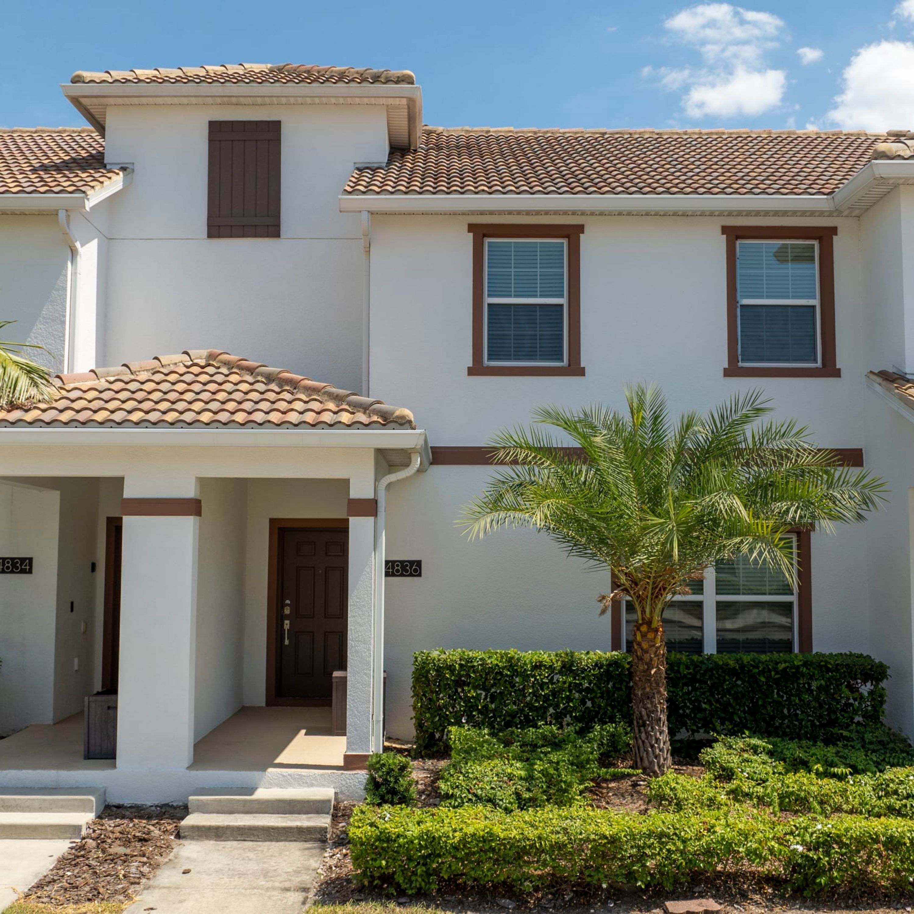 A two-story house with a tiled roof, a covered porch, and a well-maintained front yard with a palm tree and shrubs.