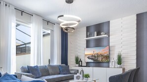 Een moderne woonkamer met een bank, salontafel en stoelen. Er is een keuken met een aanrecht en kasten. Een schilderij hangt aan de muur.